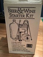 Labeled cardboard box showing Beer & Wine Starter Kit title with list of contents: glass carboy, 25L pail and lid, siphon kit, bung & air lock, sanitizing solution, hydrometer & test jar, beginner's instruction book.