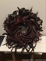 Front view of circular feather wreath in burgundy and dark hues on white surface.
