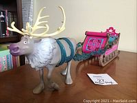 Sven figure attached to pink and blue sleigh on wooden surface, showing full figure with antlers and harness.