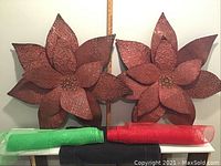 Photo showing two large burgundy metal poinsettias with embossed detailed petals and gemstone centers. Below are two rolls of metallic mesh ribbon, one red and one green.