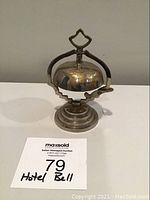 Front view of a metal hotel or desk bell with a round stepped base, floral etching in gold tone on the dome, and a decorative top handle.