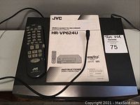 Top-down view showing black JVC VCR with original remote control and instruction manual laid on top, power cord visible.