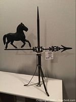 Front angle of black metal weather vane showing horse figure, vertical rod with spike, and tri-legged base.