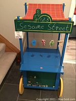 Front view showing blue wooden stand with yellow wheels, red/orange plaid fabric awning, front chalkboard with 'Sesame Street' written in white chalk and character decals, and hanging Sesame Street character decorations.