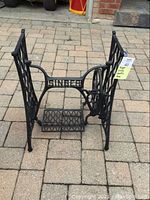Front view of the Singer wrought iron treadle legs showing 'SINGER' cast in metal, treadle pedal, and connecting wooden bar.