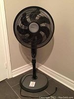 Full view of black Comfortmate standing fan against wall on tile floor, showing height and base.