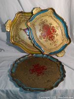 Photo of three stacked gold-finished Florentine trays showing ornate design and painted motifs in different colors.