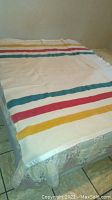 Full view of cream-colored wool blanket with horizontal stripes in red, yellow, and green spread across a mattress.