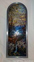 Full view of the arched stained glass window showing landscape scene with trees, waterfall, flowers and pond in multi-colored glass panel framed in metal.