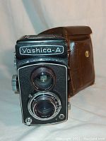 Front view of Yashica-A twin lens reflex camera showing lens and name plate with brown leather case behind it.