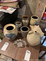 Six stoneware jugs and crocks arranged on a table with a large conch seashell and crystal clock. Variety of sizes, mostly beige with blue decoration, signs of use visible.