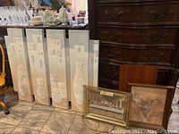 Set of 3 Mexican-themed carved panels showing stylized pottery and building facades arranged together, with the two framed pictures leaning in front on floor.