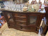 Front view of lighted buffet showing stone top, five pull-out drawers in the center, two glass-front cabinets on sides with wood framing, ornate drawer handles, and curved base.