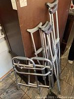 Photo showing all four items leaning against a wall on tile floor: three pairs of crutches and one folding walker