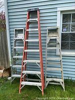 Three ladders against a house wall. Two aluminum-colored and one orange Werner ladder in the middle.
