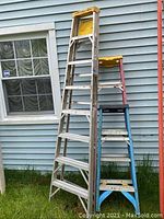 Photo showing three Werner ladders (8ft, 6ft, 4ft) outside a light blue sided building. Yellow plastic top on 8ft ladder, reddish frame on 6ft ladder, blue frame on 4ft ladder, all with paint stains and signs of wear.