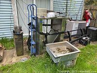 Overview photo showing Hardt rotisserie oven with fryer baskets on top and metal bowl. Atlas set and serve refrigerator/freezer visible to the right, both items outdoors on grass with signs of rust and dirt.