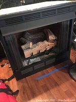 Front view of the ESA gas fireplace showing black metal housing, glass front, artificial logs inside, and vent openings on top.