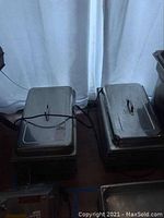 Two electric stainless steel food warmers with lids and power cords on table, natural light from window behind