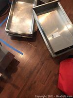 Stack of stainless steel serving pans on a wooden floor, showing two stacks with the deeper pans to the right and shallower pans to the left.