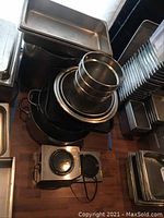Photo showing stacked large black pot and smaller stainless steel bowls and pots alongside metal and plastic trays and a two-burner hot plate on a wooden floor.