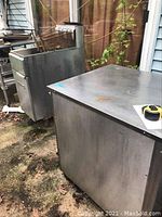 Photo of an Imperial gas deep fat fryer with fry baskets and a stainless steel holding cabinet placed outdoors showing rust and wear.