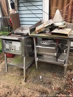 Full view of Tec infrared grill with attached wheels and adjacent stainless steel table holding pans and conical accessories.