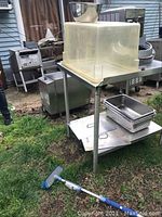 Wide shot of stainless steel table with steamer and kitchenware outdoors on grass. Shows plastic tub on top, pans on lower shelf, and metal bowl.