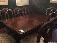 Full view of the rectangular wooden dining table with six matching chairs around it and one captain's chair visible. Dark polished wood with carved design on chair backs and light fabric seat cushions.