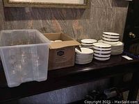 Wide view of multiple plates, bowls, cups, and stemware arranged on a dark surface showing the Mikasa Solitude china set and glasses.