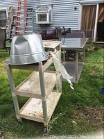 Plastic utility cart with scratches and soiling, metal basin and plastic covering draped over it, placed outdoors on grass.