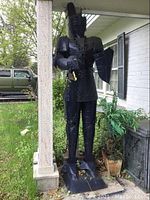 Full front side view of a navy blue painted metal suit of armor standing on a metal base next to porch column and plants.