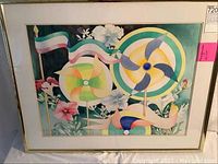 Full view of framed "Pinwheels" artwork showing colorful pinwheels, floral elements and banners, with a beige mat and gold frame.