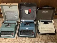 Three vintage typewriters side by side in cases on floor, left and center are blue Underwood Olivetti manual models, right is white Sears electric typewriter