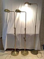 Full view of the three brass floor lamps showing adjustable height and dome-shaped lampshades with one lamp turned on.