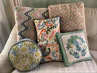 Group photo showing all five vintage needlepoint pillows on a beige couch, displaying their various colorful needlepoint designs in different shapes.