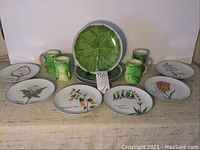 14 piece dishware lot including four large green lily pad plates stacked in the middle, surrounded by six smaller decorative botanical and animal plates, and four green frog mugs located behind the plates.