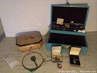 Overall view of blue velvet jewelry box open with assorted jewelry and beige carved wooden music box closed