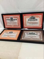 Four framed stock certificates arranged in two rows on a floor. Each has distinct colored mats and designs, showcasing vintage financial documents.