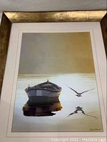 Framed print showing a boat on calm water with a bird flying, reflections visible, signed by Carlos Casamayor.