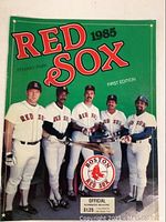 Close-up of 1985 Boston Red Sox Official Scorebook first edition cover featuring 5 players with bat, Fenway Park reference