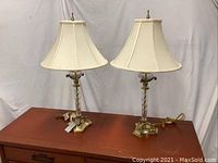 Pair of glass and brass table lamps with white bell-shaped lampshades placed on a wooden surface against a white backdrop.