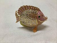 Front view of enameled fish-shaped trinket box showing gold and pink enamel with clear rhinestones and green rhinestone eye