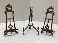 Three brass picture stands shown standing upright, showing frontal and side views.