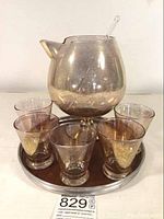 Front view of the mercury glass pitcher, cocktail glasses arranged around it, and the glass stirrer placed inside the pitcher, all on the round bar tray.