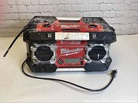 Front view of Milwaukee jobsite radio showing the speaker grills, control panel with display and buttons, and power cable attached.