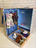 Open doll clothes trunk showing two dresses hanging inside and various pairs of doll shoes stored in the lower compartments.