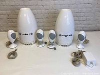 Two complete sets of JBL Encounter Alien computer speakers showing large main speakers, smaller satellite speakers, cables and power adapter on a white surface with a brick wall background.