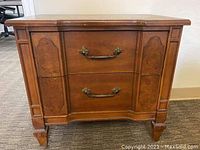 Front view of vintage wooden end table showing two drawers with decorative metal handles and carved panel details on drawer fronts and side panels.