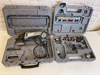 Photo showing two open gray plastic cases with molded compartments. One contains the Dremel Multi-Max tool with power cord, the other contains various accessories like sanding pads, blades, and miscellaneous attachments.